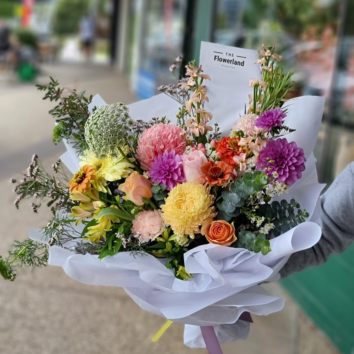 A bright artisan bouquet with orange roses, purple dahlias, and yellow chrysanthemums by The Flowerland.