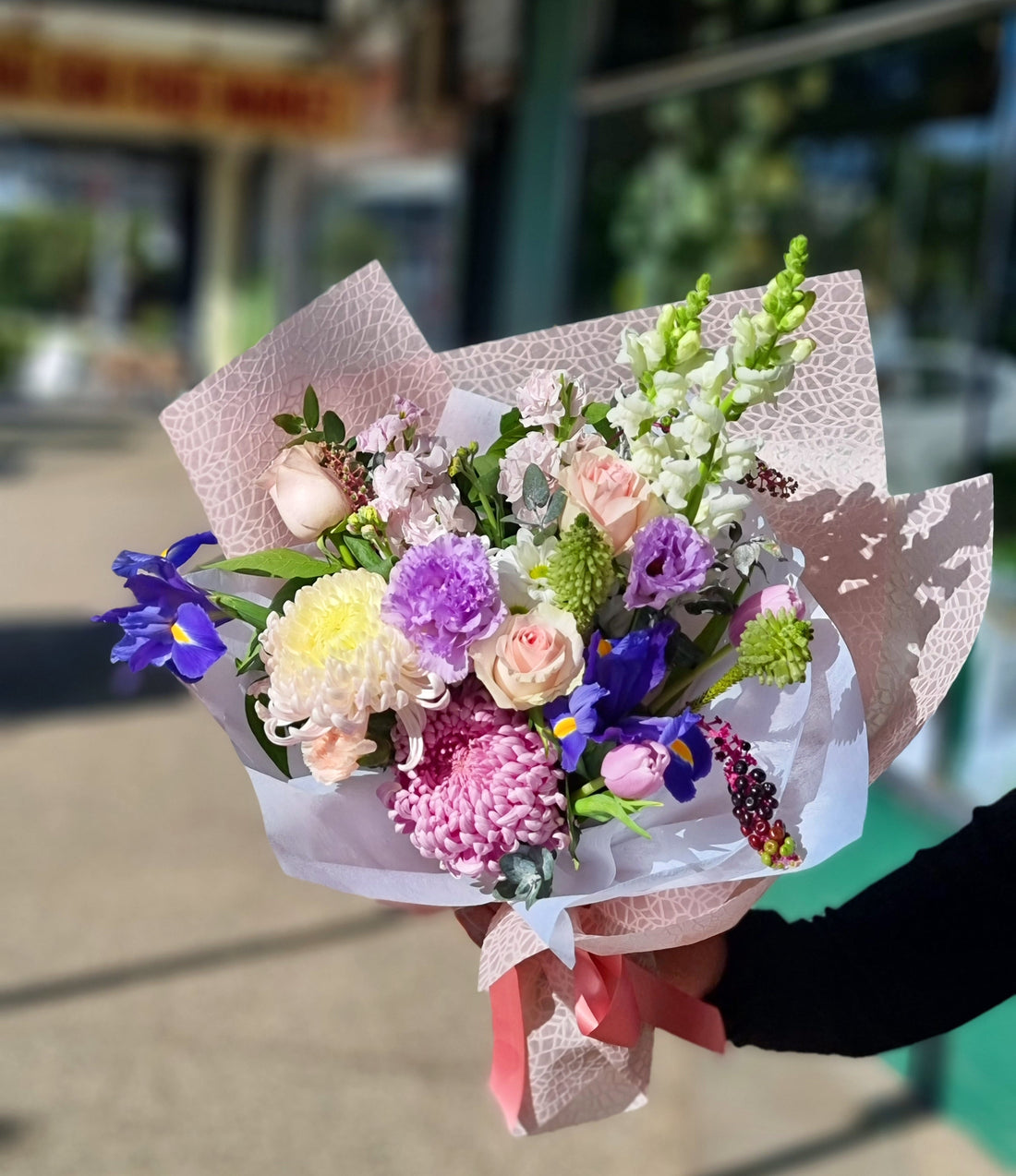 A lush floral arrangement with bright pink gerberas, deep purple irises, and soft pink roses, accented with dark foliage and held in a bright purple wrap with a matching ribbon.