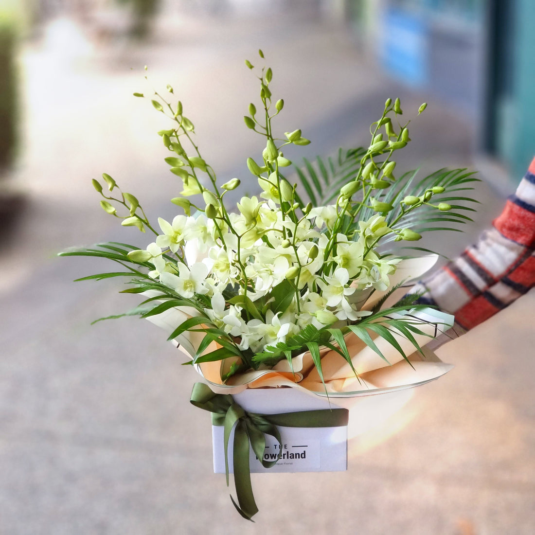 "A lush floral arrangement with a mix of pink daisies, purple irises, white protea, and red roses, held by someone in a bright blue sleeve and wrapped in patterned purple paper."