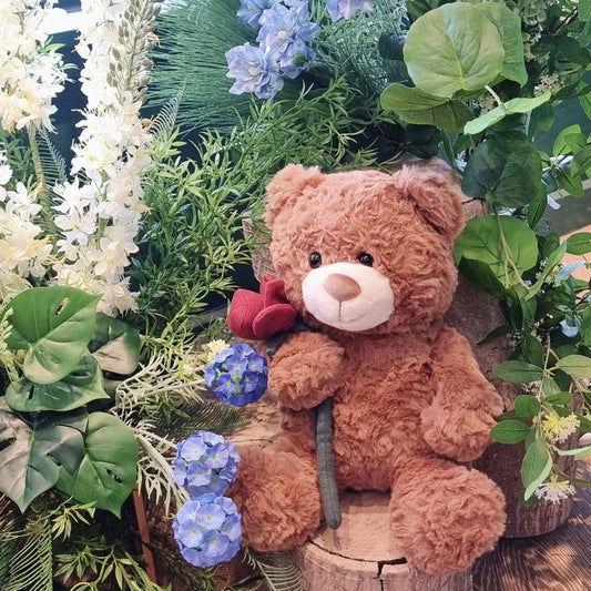 Fluffy 35cm sitting teddy bear holding a single premium red rose surrounded by garden greenery same-day delivery The Flowerland Nunawading